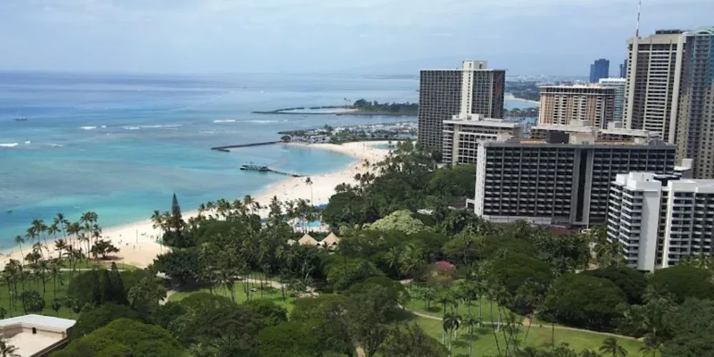 The image offers an aerial view of Fort DeRussy Beach Park and the surrounding Waikiki area. The park's lush greenery contrasts with the sandy beach and the blue ocean, while high-rise buildings frame the scene, showcasing the park's prime location in Honolulu.