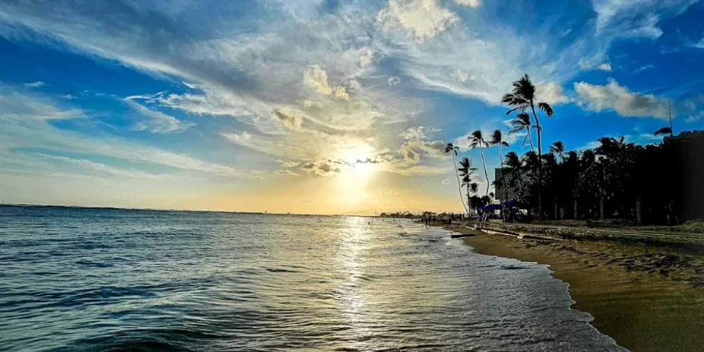 The image shows a stunning sunset at Fort DeRussy Beach Park in Waikiki, with the sun casting golden hues across the sky and ocean. Palm trees line the beach, and gentle waves lap at the shore, creating a tranquil and picturesque scene.