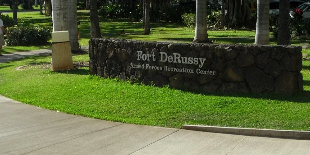 The image shows the entrance to Fort DeRussy Beach Park, marked by a stone sign that reads "Fort DeRussy Armed Forces Recreation Center." The sign is set against a backdrop of well-maintained grass and tall palm trees, inviting visitors into the park.