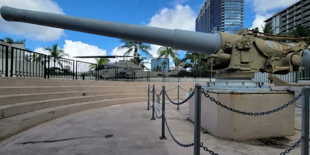 The image depicts a historic cannon on display at Fort DeRussy Beach Park. The cannon is mounted on a concrete platform surrounded by a low metal fence, with palm trees and modern buildings in the background.