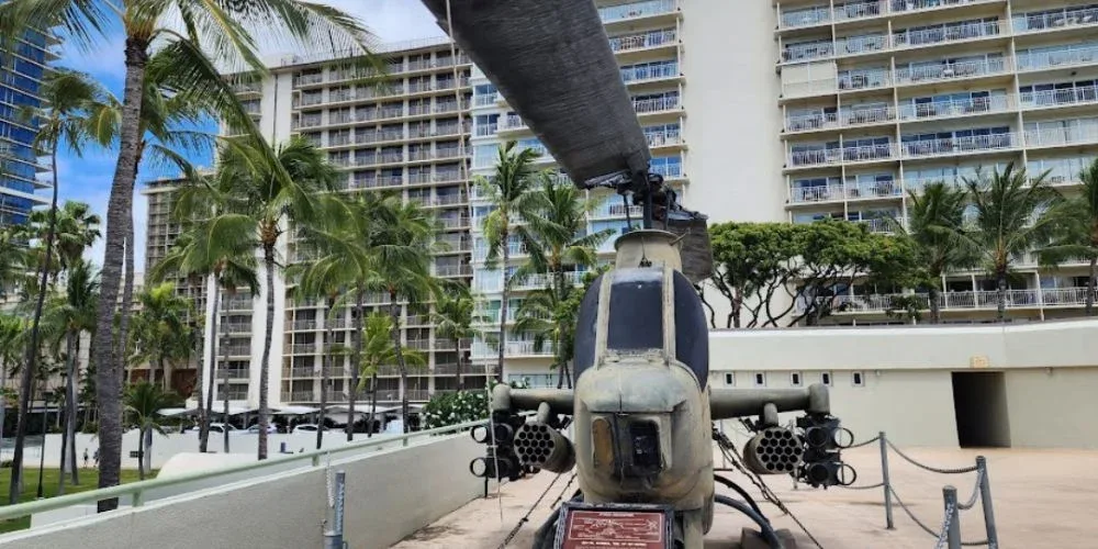 The image features a military helicopter on display at Fort DeRussy Beach Park. The helicopter is positioned in front of high-rise buildings and palm trees, illustrating the park's blend of historical military exhibits and urban surroundings.