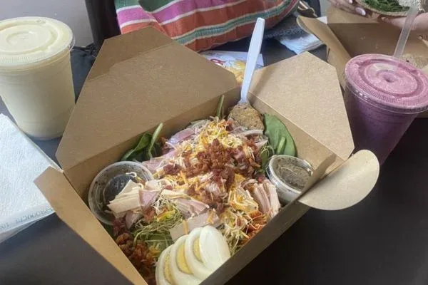 A healthy fresh salad from Pai's Deli in Waikiki, featuring mixed greens, sliced eggs, bacon bits, shredded cheese, and turkey, served in a brown takeout box with a side of dressing and accompanied by smoothies.