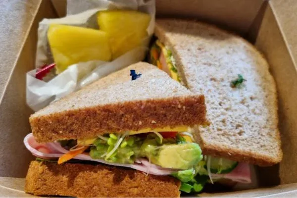 A freshly made sandwich from Pai's Deli in Waikiki, showcasing whole grain bread filled with avocado, turkey, lettuce, and other fresh vegetables, served with a side of pineapple slices in a brown takeout box.