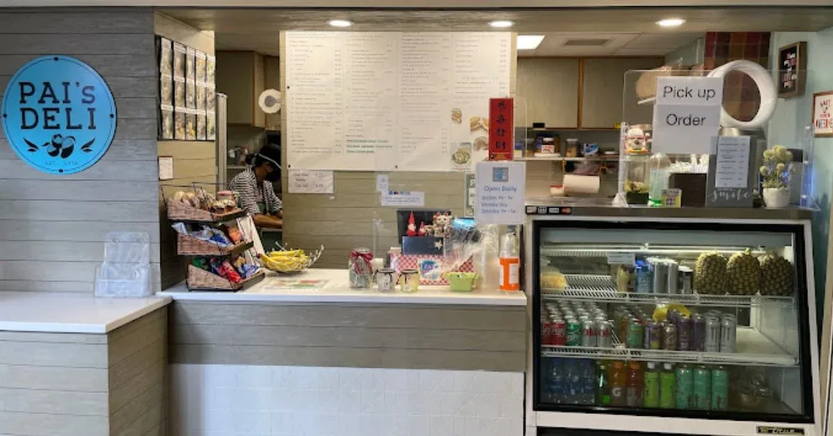 Interior view of Pai's Deli in Waikiki, showing the deli counter with a variety of snacks, beverages in a cooler, and the menu displayed on the wall behind the counter. The deli has a cozy and welcoming atmosphere, with a staff member preparing food in the background.