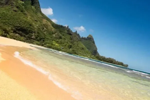 A pristine beach with clear, shallow water meeting the sandy shore, backed by lush green mountains under a bright blue sky.