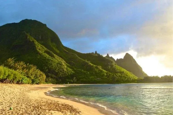 A beach scene at sunrise or sunset with the sun casting warm light on the sand and mountains in the background, creating a serene and tranquil atmosphere.