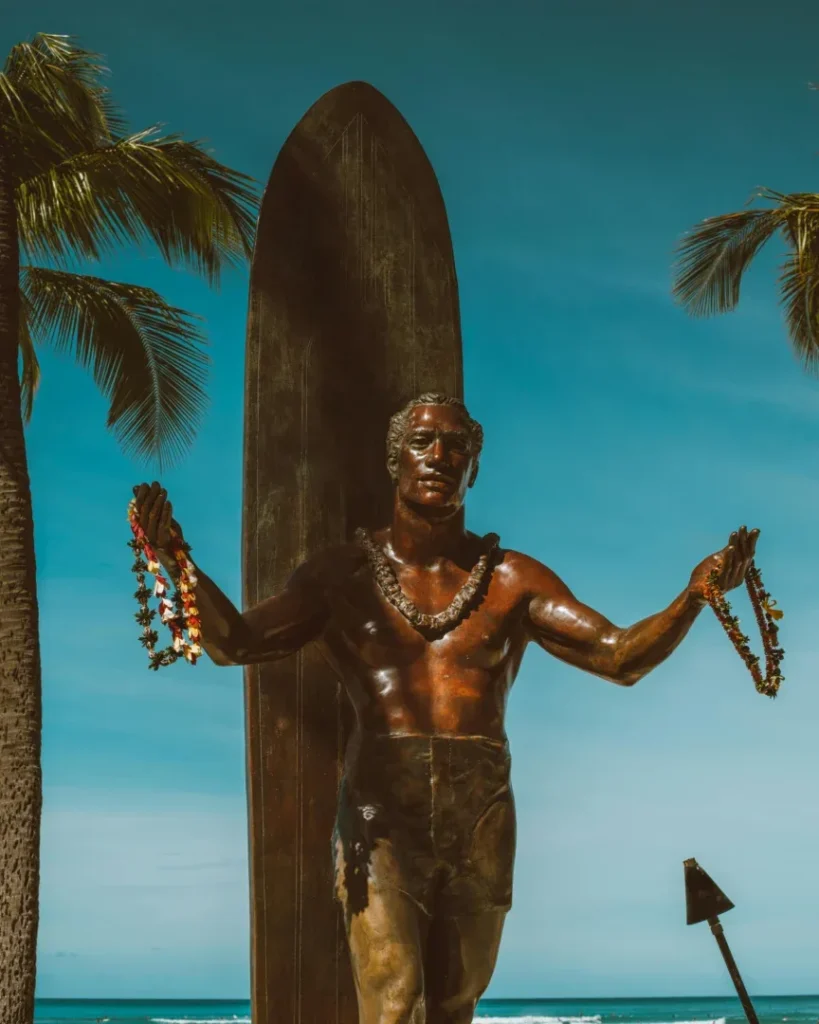 This image depicts the iconic statue of Duke Kahanamoku, a legendary Hawaiian surfer and Olympic swimmer, located at Kalakaua Avenue, Waikiki Beach, Honolulu, HI 96815, United States. The bronze statue shows Duke with outstretched arms, wearing a lei around his neck, with a large surfboard standing upright behind him. Palm trees and a clear blue sky provide a picturesque background, highlighting the tropical and serene setting.