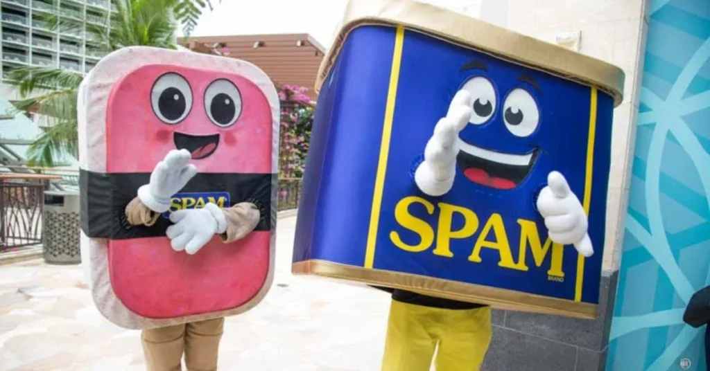 Two people dressed in mascot costumes at a Spam Jam event in Waikiki, 2017. One costume resembles a can of Spam, with a blue and yellow design and a smiling face. The other resembles a slice of Spam, with a pink and black design and a smiling face. They are posing and waving, with palm trees and buildings visible in the background.